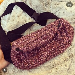 May include: A handmade fanny pack in shades of pink and purple, with a black adjustable strap and buckle. The bag is made of a thick, textured yarn, and has a zippered closure. The strap is made of a durable black fabric.
