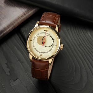 Könnte beinhalten: Eine goldfarbene Armbanduhr mit braunem Lederarmband. Das cremefarbene Zifferblatt hat ein einzigartiges Design mit einem roten Sekundenzeiger und einem kleinen goldfarbenen Zifferblatt. Das Armband hat ein Krokodilmuster.