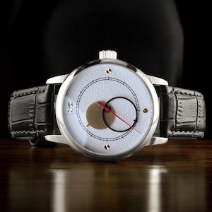 Op de afbeelding: Een zilveren polshorloge met een witte wijzerplaat, zwarte en gouden accenten en een rode secondewijzer. Het horloge heeft een zwarte leren band. Het horloge wordt gepresenteerd op een houten oppervlak tegen een donkere achtergrond.