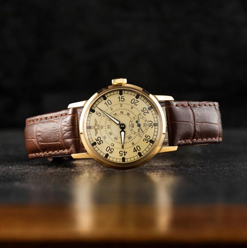 May include: A gold-toned wristwatch with a cream-colored face and black numerals. The watch features a brown leather band with a crocodile pattern. The watch hands are black, and the watch is set against a dark background.