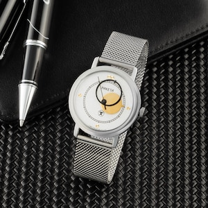 May include: A silver-tone wristwatch with a white dial, a gold-colored circular accent, and a mesh metal strap. The dial features the text "PAKETA" in black, thin black hands, and small gold-tone markers. The watch has a round case and a crown.
