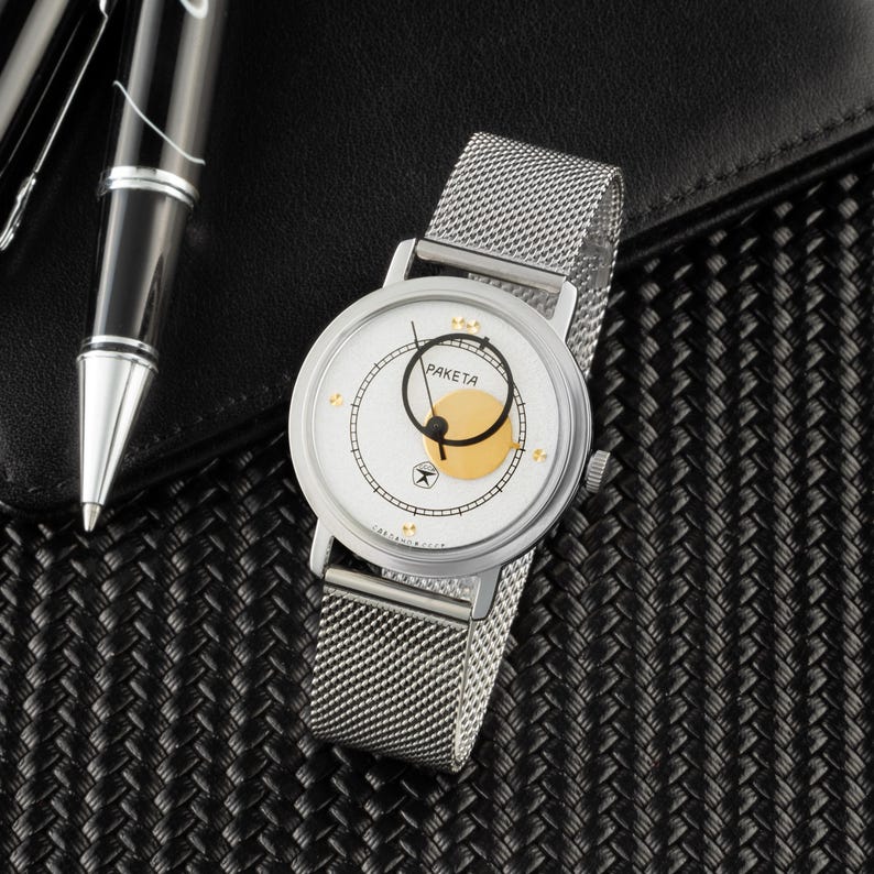 May include: A silver-tone wristwatch with a white dial, a gold-colored circular accent, and a mesh metal strap. The dial features the text "PAKETA" in black, thin black hands, and small gold-tone markers. The watch has a round case and a crown.