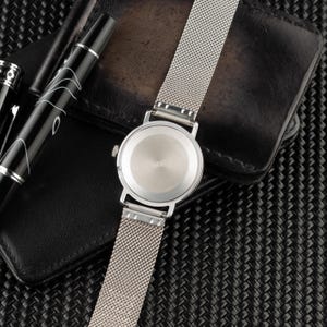May include: A silver-toned wristwatch with a circular case and a fine mesh metal strap. The back of the watch case is smooth and metallic, featuring a small winding crown on the side. The watch is positioned alongside a black fountain pen.