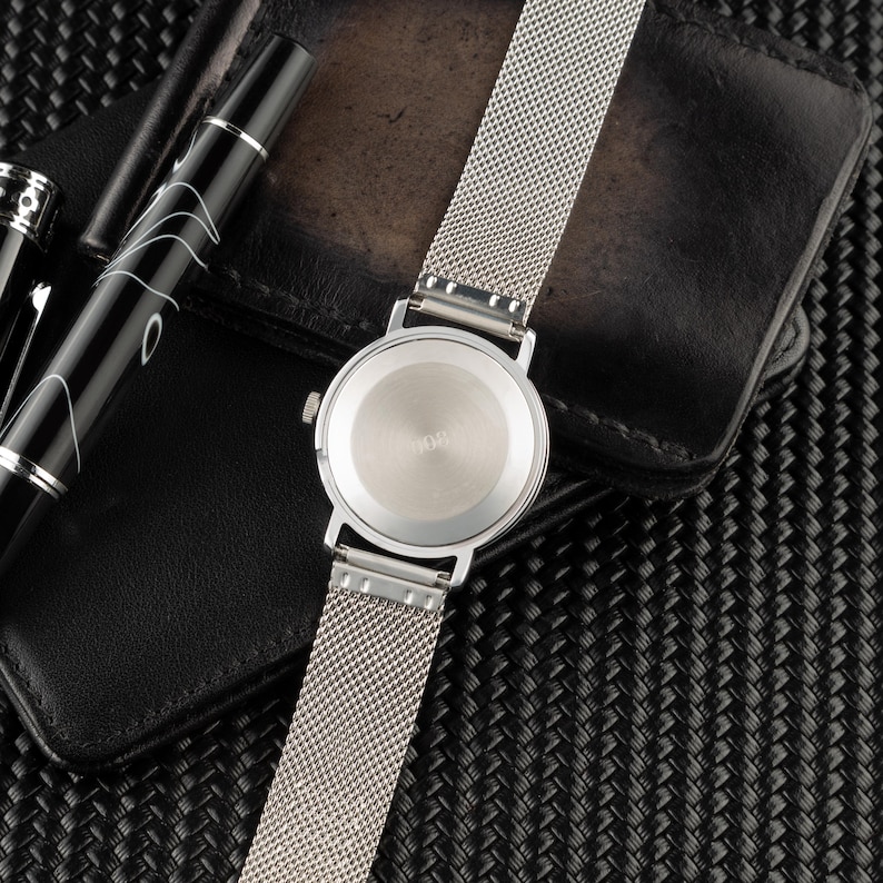 May include: A silver-toned wristwatch with a circular case and a fine mesh metal strap. The back of the watch case is smooth and metallic, featuring a small winding crown on the side. The watch is positioned alongside a black fountain pen.