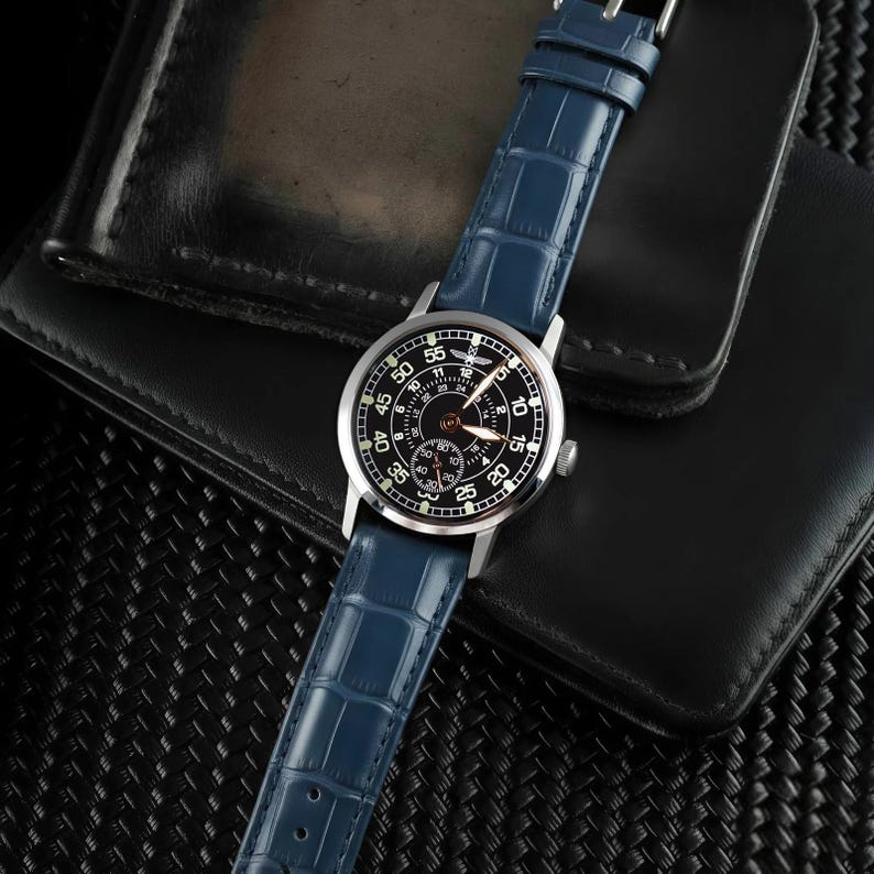May include: A silver-cased wristwatch with a black face and a blue leather band. The watch face displays white numerals and hands. The blue band has a crocodile pattern. The watch is set against a black leather wallet.