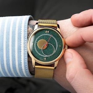 May include: A gold-toned wristwatch with a green face and a gold mesh band. The watch face features a gold circle and a red hand. The watch is being adjusted by a person wearing a blue and white striped shirt.
