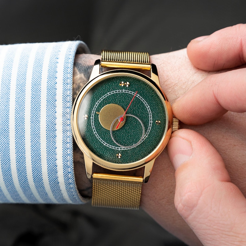 May include: A gold-toned wristwatch with a green face and a gold mesh band. The watch face features a gold circle and a red hand. The watch is being adjusted by a person wearing a blue and white striped shirt.