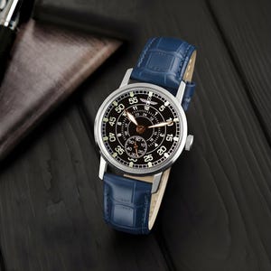 May include: A silver-cased wristwatch with a black face and a blue leather band. The watch face features white numerals and hands, with a smaller subdial. The band has a crocodile pattern. The watch is displayed on a dark wooden surface.