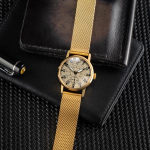 May include: A gold-toned wristwatch with a mesh band and a cream-colored face. The watch features black numerals and hands, with a smaller dial at the bottom. The watch is set on a black leather wallet.
