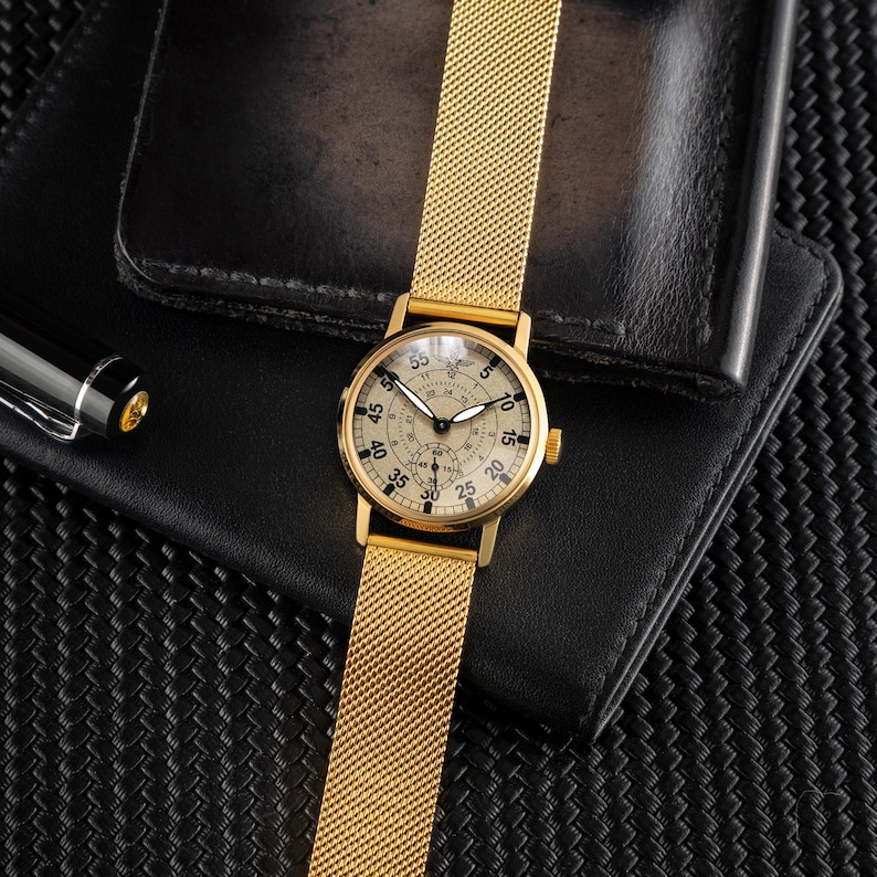 May include: A gold-toned wristwatch with a mesh band and a cream-colored face. The watch features black numerals and hands, with a smaller dial at the bottom. The watch is set on a black leather wallet.