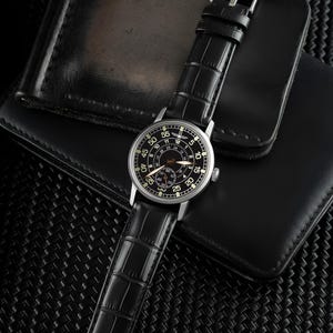 May include: A black wristwatch with a silver case and a black leather strap with a crocodile pattern. The watch face is black with white numerals and hands. The watch is on a black leather wallet.