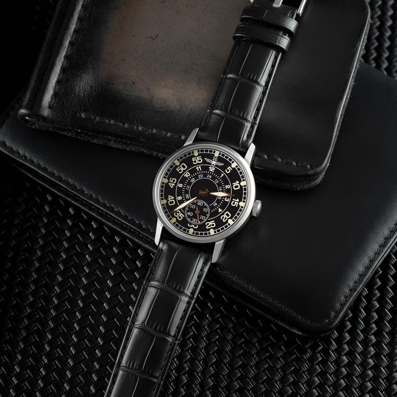 May include: A black wristwatch with a silver case and a black leather strap with a crocodile pattern. The watch face is black with white numerals and hands. The watch is on a black leather wallet.