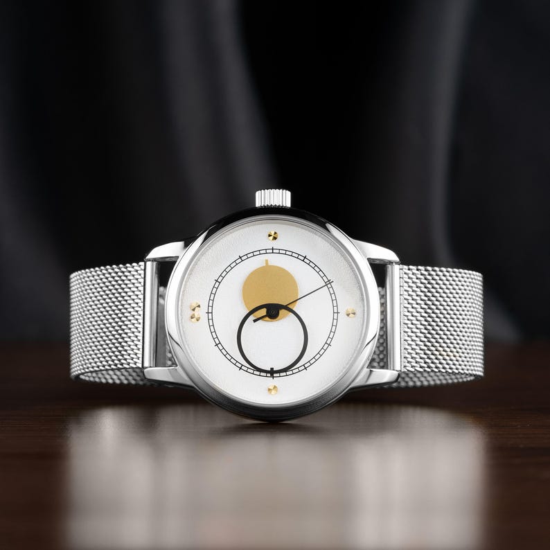 K&ouml;nnte beinhalten: Eine silberne Armbanduhr mit wei&szlig;em Zifferblatt, das einen goldfarbenen Kreis und einen schwarzen Kreis aufweist. Die Uhr hat ein Mesh-Armband und ein silbernes Geh&auml;use. Die Uhr wird auf einer dunklen Holzoberfl&auml;che pr&auml;sentiert.