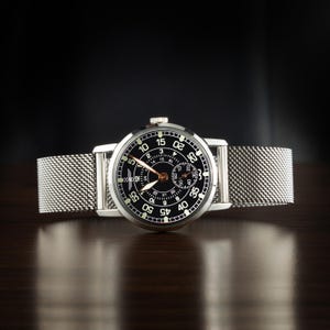 May include: A silver wristwatch with a black face and a mesh band. The watch face features white numerals and hands, with a smaller dial. The watch is displayed on a dark wooden surface.