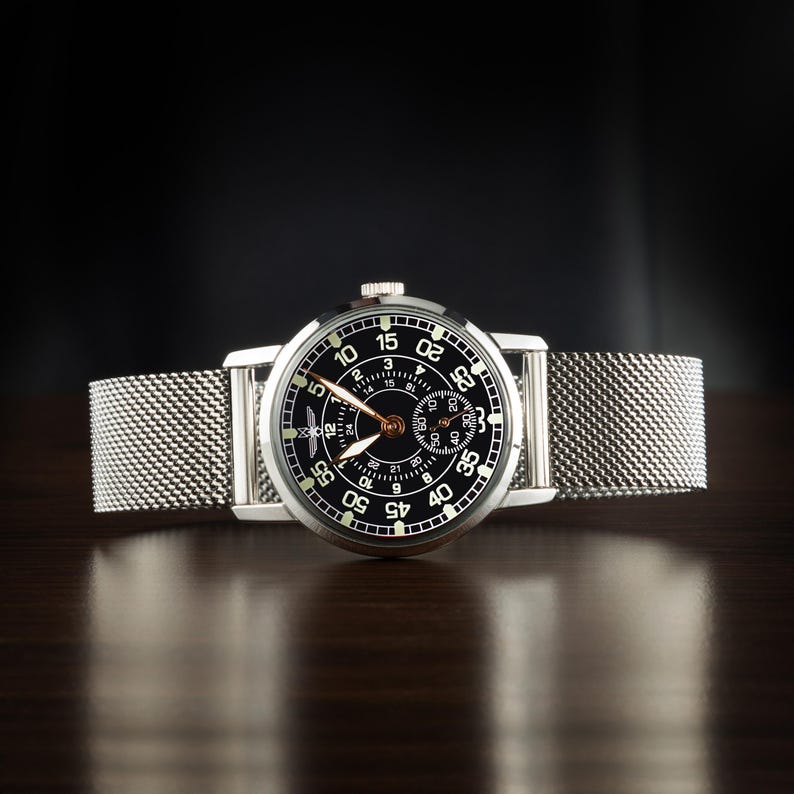 May include: A silver wristwatch with a black face and a mesh band. The watch face features white numerals and hands, with a smaller dial. The watch is displayed on a dark wooden surface.