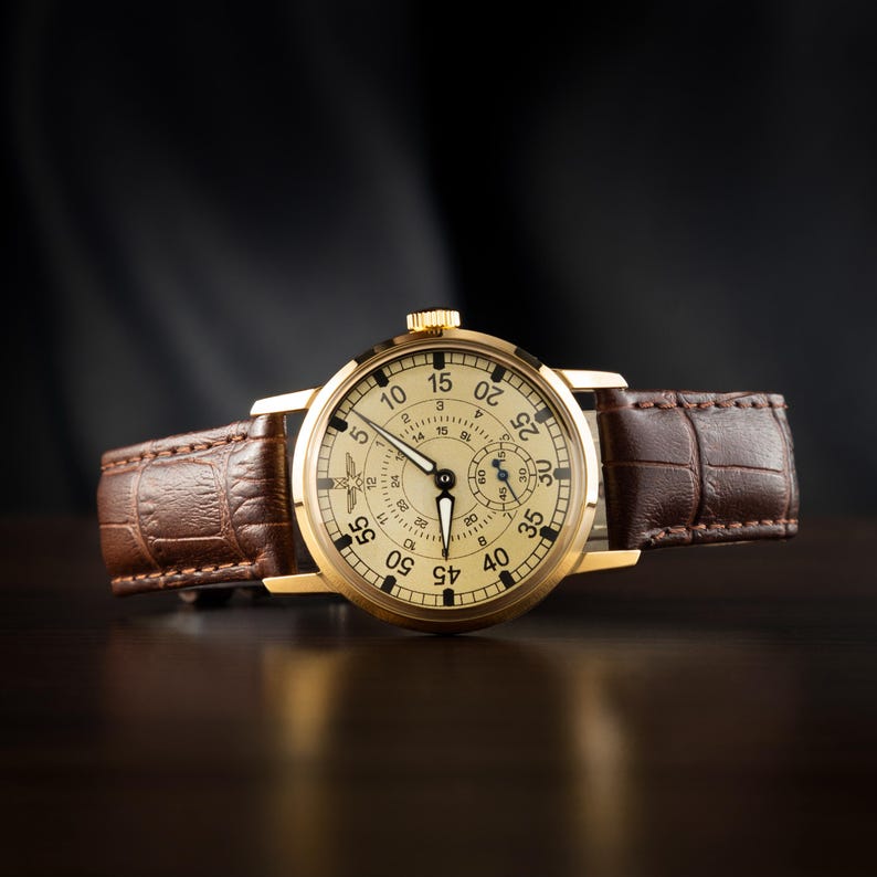 May include: A gold-toned wristwatch with a cream-colored face and black numerals. The watch features a brown leather band with a crocodile pattern. The watch hands are black, and the watch is set against a dark background.