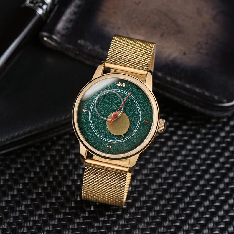May include: Gold-toned wristwatch with a green face and a mesh band. The watch face features a circular design with a red hand and gold accents. The watch is displayed on a textured black surface.