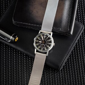 May include: A silver wristwatch with a black face and a mesh band is displayed. The watch is set against a black leather wallet and a black pen. The watch face features white numerals and hands, with a smaller dial at the bottom.