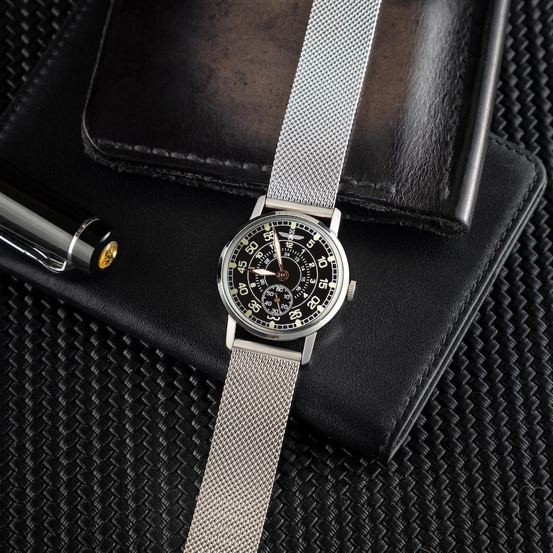 May include: A silver wristwatch with a black face and a mesh band is displayed. The watch is set against a black leather wallet and a black pen. The watch face features white numerals and hands, with a smaller dial at the bottom.