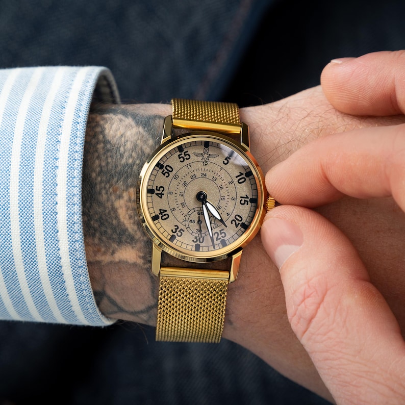 May include: A gold-toned wristwatch with a mesh band and a round face. The watch face has black numerals and hands against a light-colored background. The watch is being adjusted by a person wearing a blue and white striped shirt.