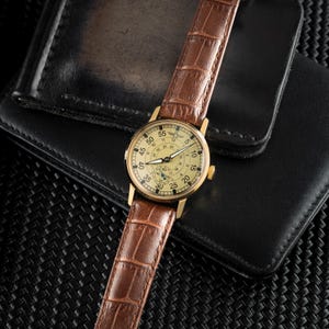 May include: A gold-toned wristwatch with a cream-colored face and brown leather band. The watch features a detailed dial with multiple sub-dials and a brown, textured leather strap. The watch is resting on a black leather wallet.