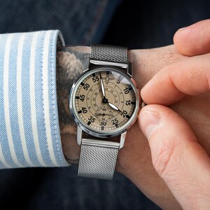 May include: A silver wristwatch with a mesh band and a beige face. The watch has black hands and numerals, with a smaller dial at the bottom. The watch is being adjusted by a person wearing a blue and white striped shirt.
