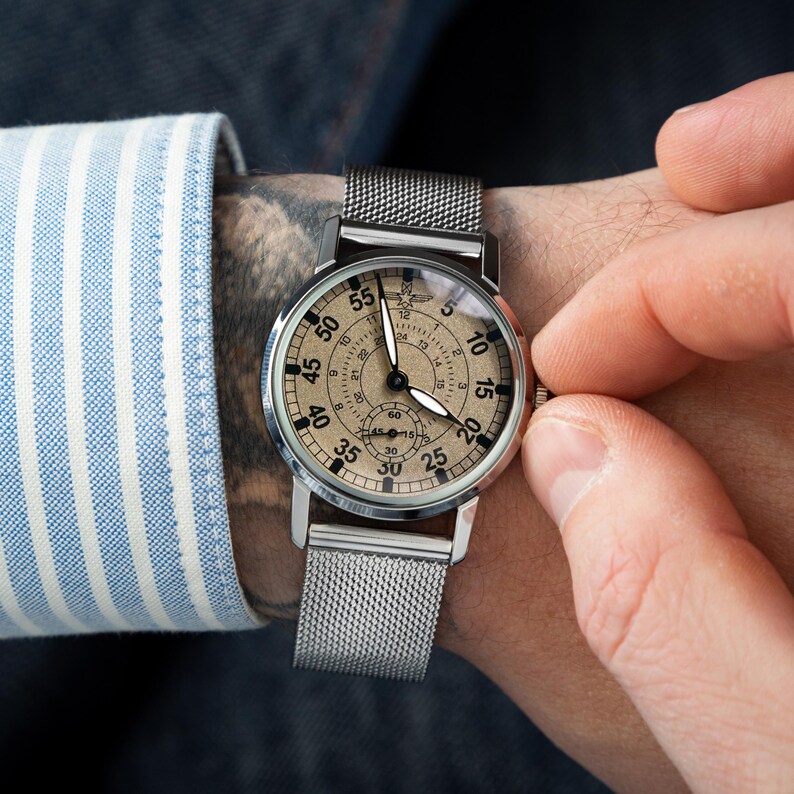 May include: A silver wristwatch with a mesh band and a beige face. The watch has black hands and numerals, with a smaller dial at the bottom. The watch is being adjusted by a person wearing a blue and white striped shirt.