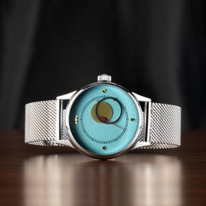 May include: A silver wristwatch with a light blue face and a mesh band. The watch face features a gold and black design with a red hand. The watch has a round face and a silver case. The band is made of a fine, woven metal.