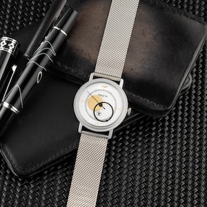 May include: A silver-tone wristwatch with a white dial, a gold circular accent, and a black circular hand. The watch features the text PAKETA on the face and includes a silver mesh metal strap. The design includes small gold markers at the hour positions.
