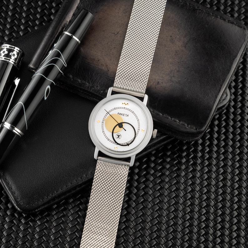 May include: A silver-tone wristwatch with a white dial, a gold circular accent, and a black circular hand. The watch features the text PAKETA on the face and includes a silver mesh metal strap. The design includes small gold markers at the hour positions.