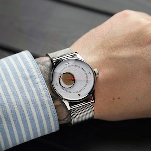 K&ouml;nnte beinhalten: Eine silberne Armbanduhr mit wei&szlig;em Zifferblatt und einzigartigem Design, einschlie&szlig;lich eines goldfarbenen Kreises und eines roten Zeigers. Die Uhr hat ein Mesh-Armband und wird am Handgelenk getragen. Die Person tr&auml;gt ein blau-wei&szlig; gestreiftes Hemd.