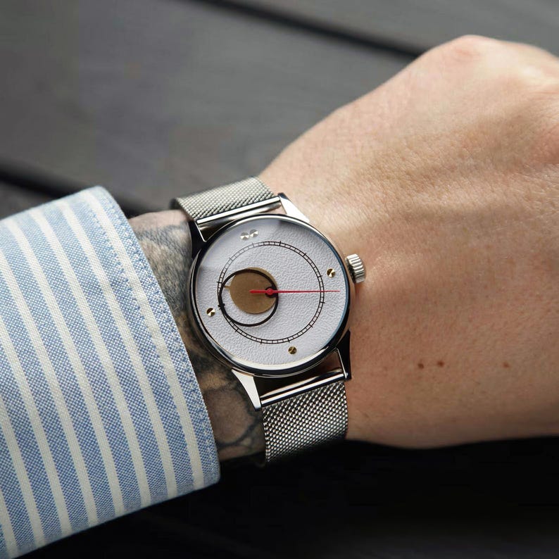 K&ouml;nnte beinhalten: Eine silberne Armbanduhr mit wei&szlig;em Zifferblatt und einzigartigem Design, einschlie&szlig;lich eines goldfarbenen Kreises und eines roten Zeigers. Die Uhr hat ein Mesh-Armband und wird am Handgelenk getragen. Die Person tr&auml;gt ein blau-wei&szlig; gestreiftes Hemd.