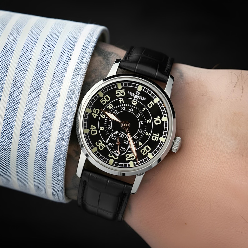 May include: A black wristwatch with a silver case and a black leather strap. The watch face features white numerals and hands, with a smaller dial. The watch is on a wrist, next to a blue and white striped shirt sleeve.