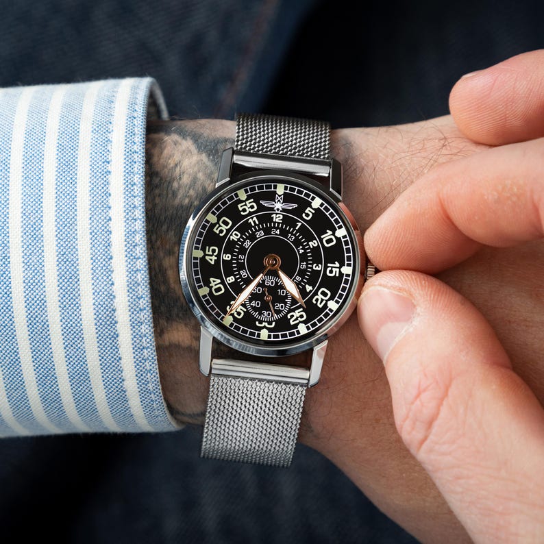 May include: A black and silver wristwatch with a mesh band. The watch face is black with white numerals and hands. The watch is being adjusted by a person wearing a blue and white striped shirt.