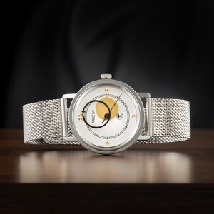May include: A silver-tone wristwatch with a mesh metal strap and a round white dial. The dial features a gold circle, a black circular hand, thin black hour markers, and the text PAKETA. A small crown is positioned at the top of the watch case.