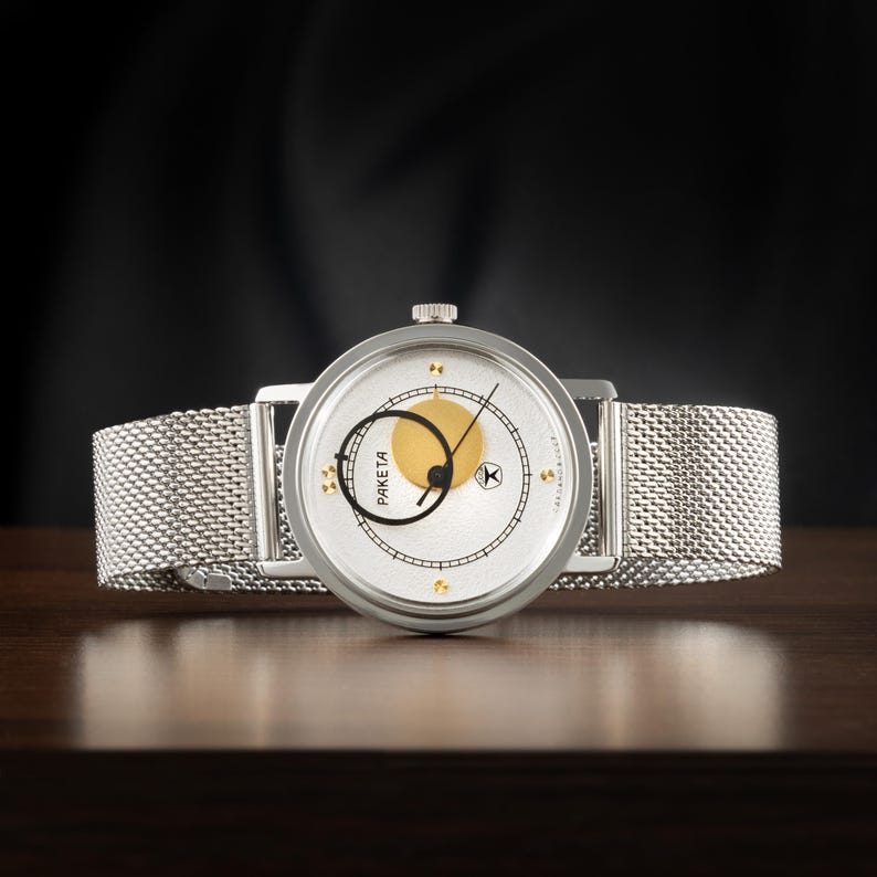 May include: A silver-tone wristwatch with a mesh metal strap and a round white dial. The dial features a gold circle, a black circular hand, thin black hour markers, and the text PAKETA. A small crown is positioned at the top of the watch case.
