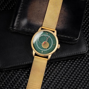 May include: A gold-toned wristwatch with a green face and gold mesh band. The watch face features a unique design with a red hand and a gold-colored accent. The watch is set against a black leather wallet.