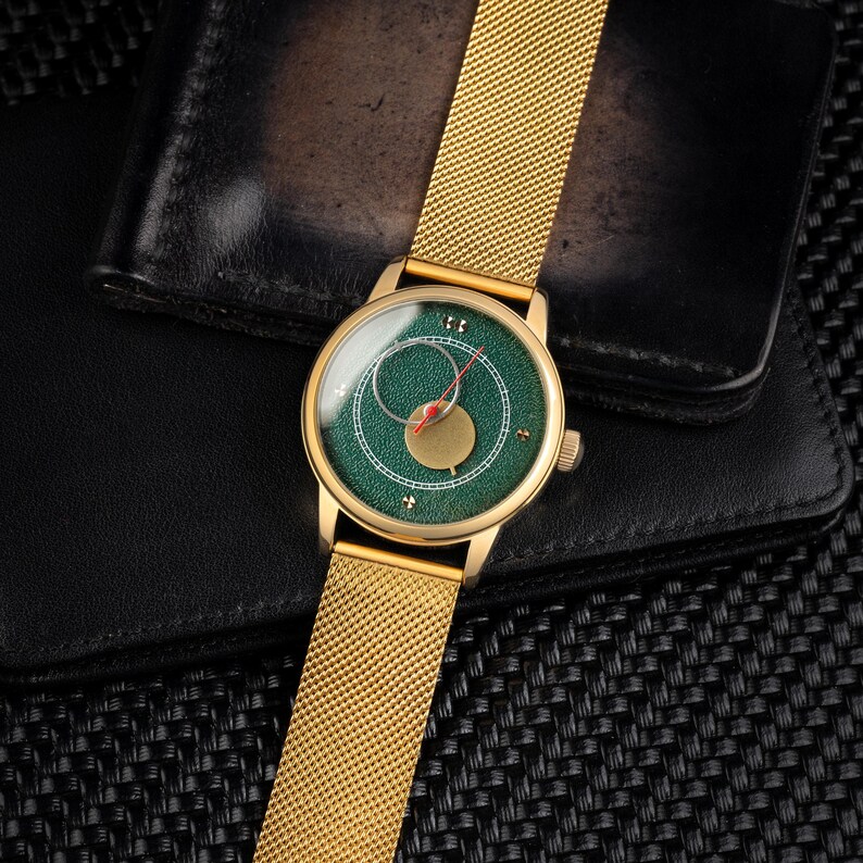 May include: A gold-toned wristwatch with a green face and gold mesh band. The watch face features a unique design with a red hand and a gold-colored accent. The watch is set against a black leather wallet.