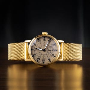 May include: A gold-toned wristwatch with a mesh band. The watch face is beige with black numerals and hands. The watch has a small seconds dial and a crown. The band is a woven mesh design.