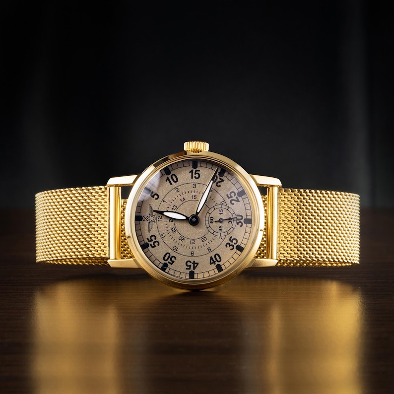 May include: A gold-toned wristwatch with a mesh band. The watch face is beige with black numerals and hands. The watch has a small seconds dial and a crown. The band is a woven mesh design.