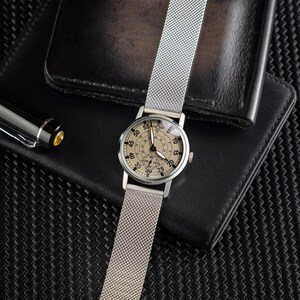 May include: A silver wristwatch with a mesh band, featuring a beige face with black numerals and hands. The watch is displayed on a black leather wallet, with a pen visible in the background. The watch face has a diameter of approximately 1.5 inches.