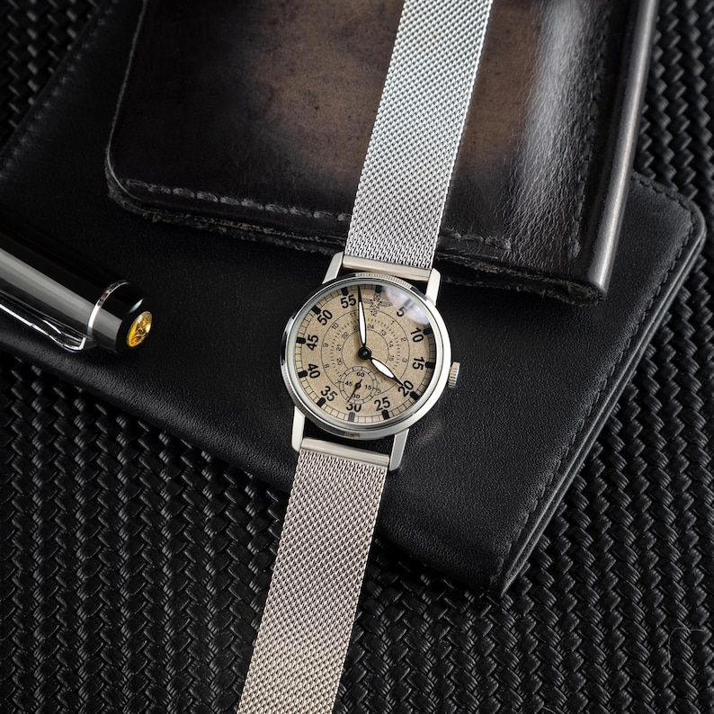 May include: A silver wristwatch with a mesh band, featuring a beige face with black numerals and hands. The watch is displayed on a black leather wallet, with a pen visible in the background. The watch face has a diameter of approximately 1.5 inches.