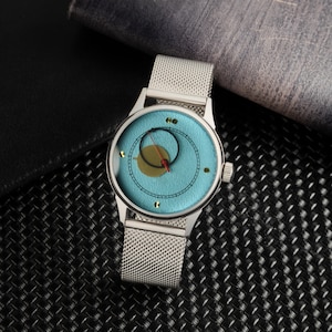 May include: A silver wristwatch with a light blue face featuring a gold circle and a red hand. The watch has a mesh band and is set against a dark background.