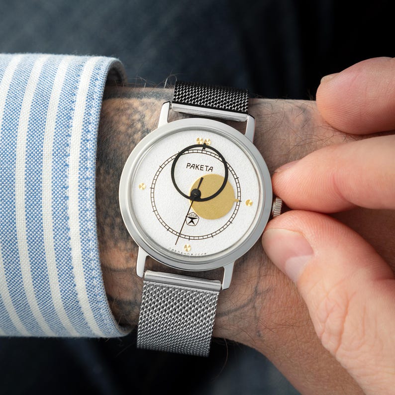 May include: A silver-tone wristwatch with a white textured dial, featuring the text PAKETA, a black circular frame, and a gold-tone circular accent. The watch has a silver-tone mesh strap, a thin black hand, and small gold-tone markers on the dial.