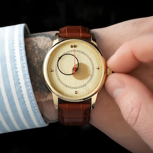 May include: A gold-toned wristwatch with a brown leather band. The watch face features a unique design with a red second hand and a black circular pattern. The watch is being adjusted by a person wearing a striped blue and white shirt.