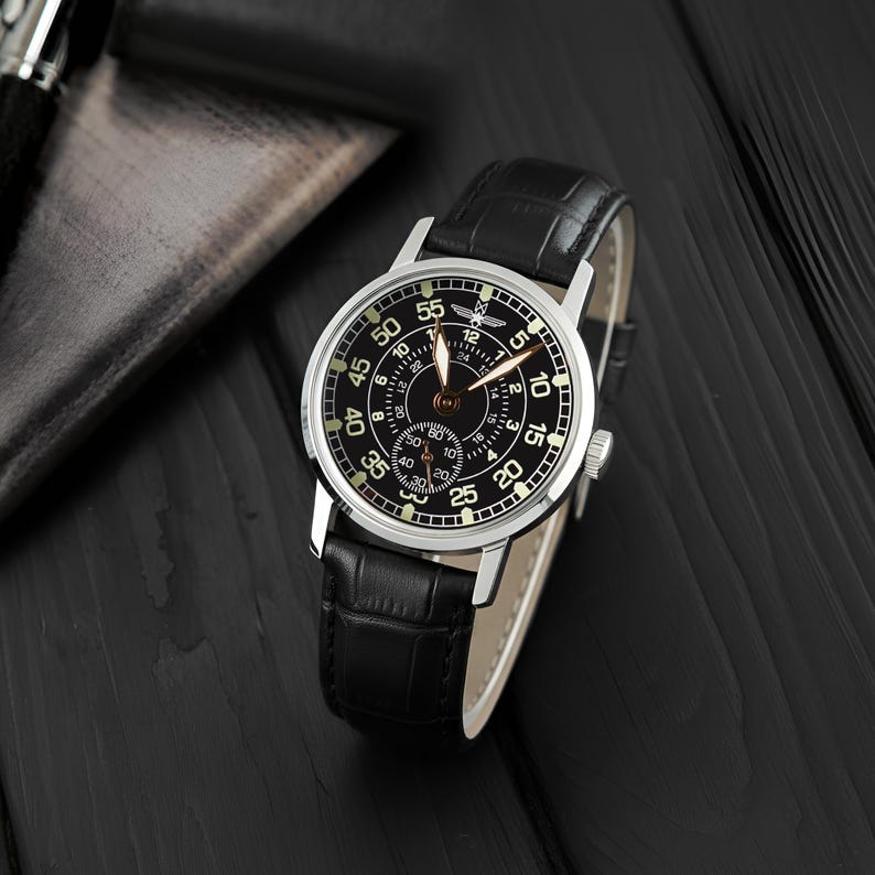 May include: A black wristwatch with a silver case and a black leather strap. The watch face is black with white numerals and hands. The watch is displayed on a dark wooden surface.