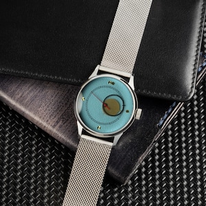 May include: A silver wristwatch with a light blue face and a mesh band. The watch face features a circular design with a gold-colored center and red hands. The watch is set against a black leather wallet and a textured background.