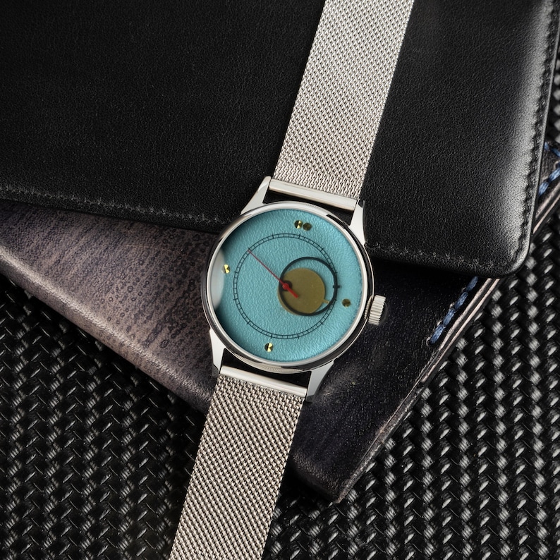 May include: A silver wristwatch with a light blue face and a mesh band. The watch face features a circular design with a gold-colored center and red hands. The watch is set against a black leather wallet and a textured background.