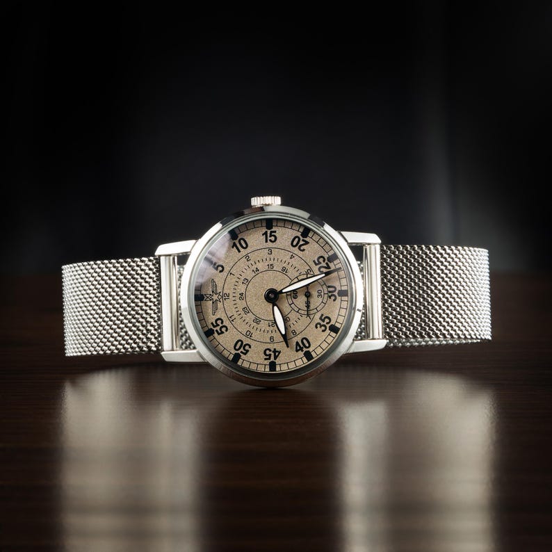 May include: A silver wristwatch with a mesh band. The watch face is beige with black numerals and hands. The watch has a diameter of approximately 1.5 inches. The watch is displayed on a dark wooden surface.