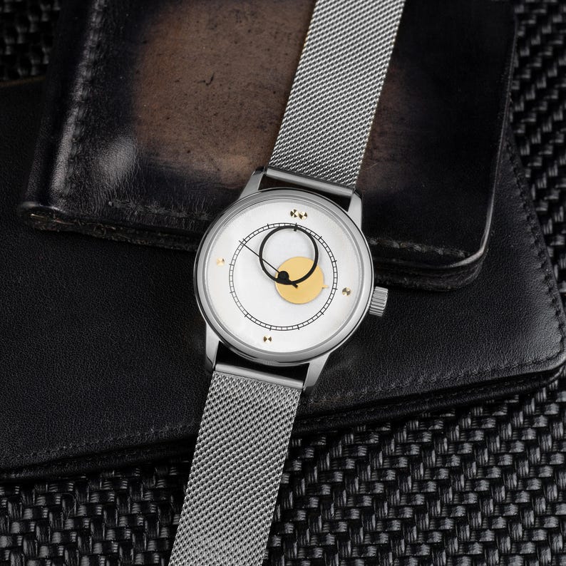 K&ouml;nnte beinhalten: Eine silberne Armbanduhr mit wei&szlig;em Zifferblatt und einem goldfarbenen Kreisdesign. Die Uhr hat ein Mesh-Armband und liegt auf einem schwarzen Lederportemonnaie und einer strukturierten schwarzen Oberfl&auml;che.
